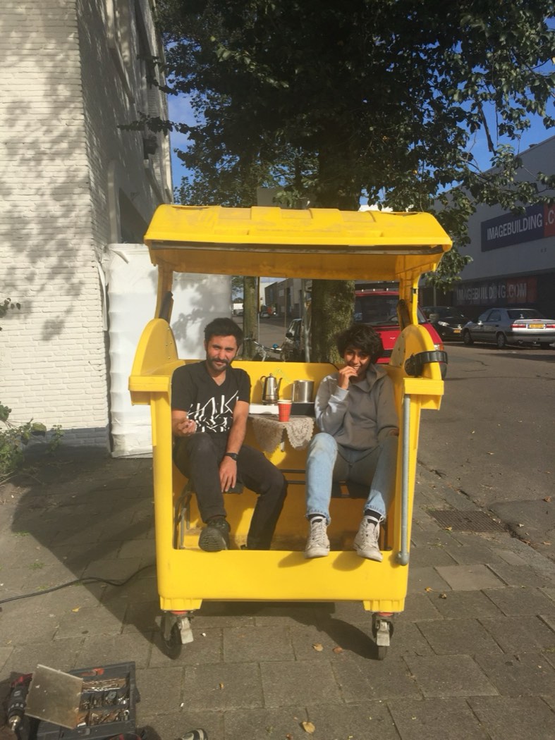 Two people having coffee inside a converted wheelie bin, roof up, parked on the street
